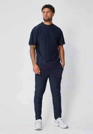 Homme avec des tatouages portant un t-shirt bleu marine et un pantalon, debout avec une main dans la poche, portant des baskets blanches sur un fond uni.