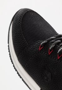 Baskets noires avec dessus en cuir texturé et mesh, accents rouges sur les lacets, et une semelle intermédiaire blanche. Gros plan montrant les détails du matériau et du design.
