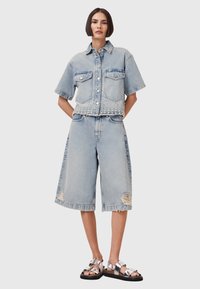 Giacca in denim azzurro chiaro a maniche corte e chiusura con bottoni, abbinata a pantaloni corti larghi che arrivano al ginocchio con bordi sfilacciati.