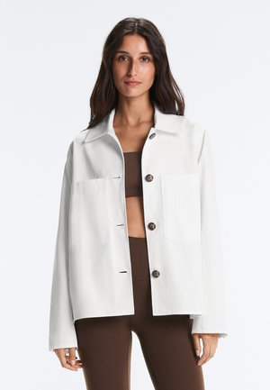 Donna con capelli lunghi e scuri che indossa una giacca bianca con bottoni sopra un crop top marrone e pantaloni marroni a vita alta, in piedi davanti a uno sfondo bianco.
