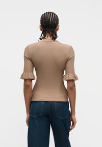 Personne aux cheveux tressés portant un haut à manches courtes côtelé marron clair avec des poignets à volants et un jean en denim bleu foncé, vue de dos.