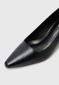 Zapatos de tacón delgado de cuero negro con punta afilada y textura suave, presentando un corte bajo en la parte delantera, con detalles de costura mínimos y una silueta estilizada.