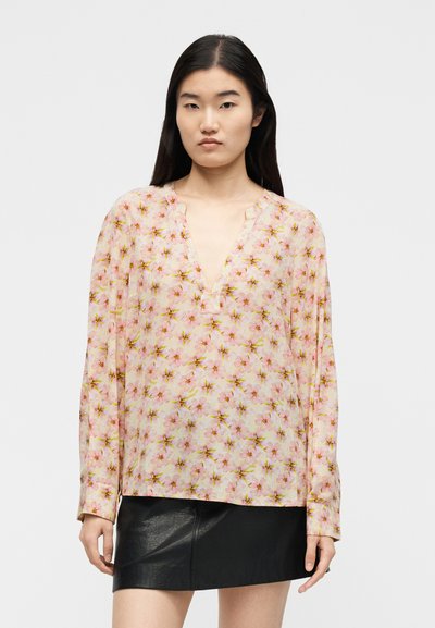Jeune femme aux longs cheveux foncés portant un chemisier transparent rose à motif floral avec un col en V et une mini-jupe en cuir noir sur un fond blanc.