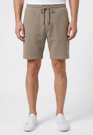 Man draagt beige shorts met trekkoord en gestructureerde stof, wit T-shirt en witte sneakers, staand tegen een effen witte achtergrond.