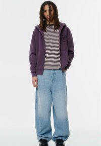 Felpa viola con zip e logo sul petto, indossata sopra una maglietta a righe. Jeans larghi di colore blu chiaro e scarpe nere completano il look.