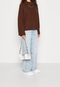 Suéter marrón con acanalado y cuello alto, combinado con jeans de pierna ancha en azul claro. Bolso blanco con detalles negros y zapatos blancos sin cordones.