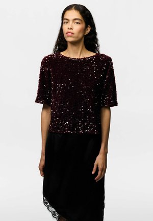 Femme aux longs cheveux bouclés foncés, portant un top bordeaux pailleté à manches courtes et une jupe noire avec un bord en dentelle, debout devant un fond uni.