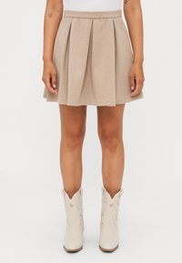 Falda plisada beige hecha de tejido texturizado, con una cintura ajustada y un dobladillo acampanado. Llevada con botas vaqueras blancas.