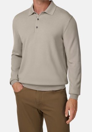 Beiger Langarm-Polo-Pullover mit gerippten Bündchen und Saum, kombiniert mit braunen Hosen, getragen von einer stehenden Person mit einer Hand in der Tasche.