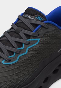 Μαύρο και μπλε αθλητικό παπούτσι Skechers με γκρι κορδόνια, υφή στην επάνω επιφάνεια, λογότυπο στη γλώσσα και σχεδιασμό σόλας με αερισμό.