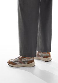 Persona che indossa pantaloni larghi grigi e sneakers grigie, marroni e arancioni, in piedi su una superficie bianca.