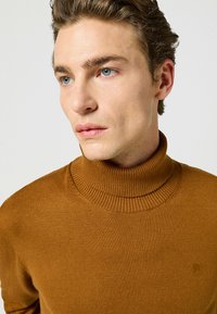 Brązowy sweter z golfem o prążkowanej strukturze i kołnierzu, z delikatnym detalem logo na piersi, wykonany z miękkiego materiału.