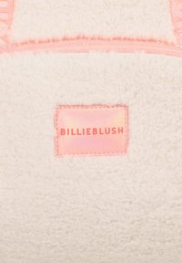 Chlupatá krémová tkanina s jemnou strukturou, opatřená lesklou růžovou etiketou s nápisem "BILLIEBLUSH." Ozdobena ladící růžovou lemovkou.