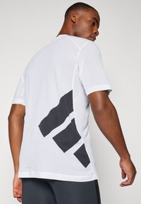 Chemise à manches courtes pour homme, en matériau en maille blanche avec un motif géométrique noir au dos. Texture lisse, col rond, coupe athlétique.