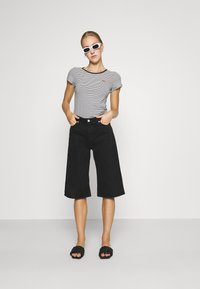 Top cu mânecă scurtă în dungi, cu culote din denim negre, sandale negre cu talpă plată și ochelari de soare albi. Fundal simplu, design minimalist.