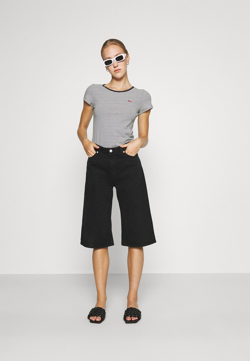 Top cu mânecă scurtă în dungi, cu culote din denim negre, sandale negre cu talpă plată și ochelari de soare albi. Fundal simplu, design minimalist.