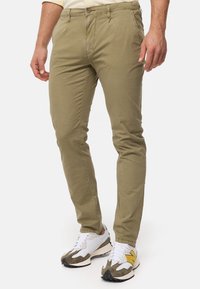 Chinos kaki ajustés, fabriqués en coton léger. Présentent une fermeture à bouton à l'avant et des poches latérales. Associés à des baskets blanches et grises.