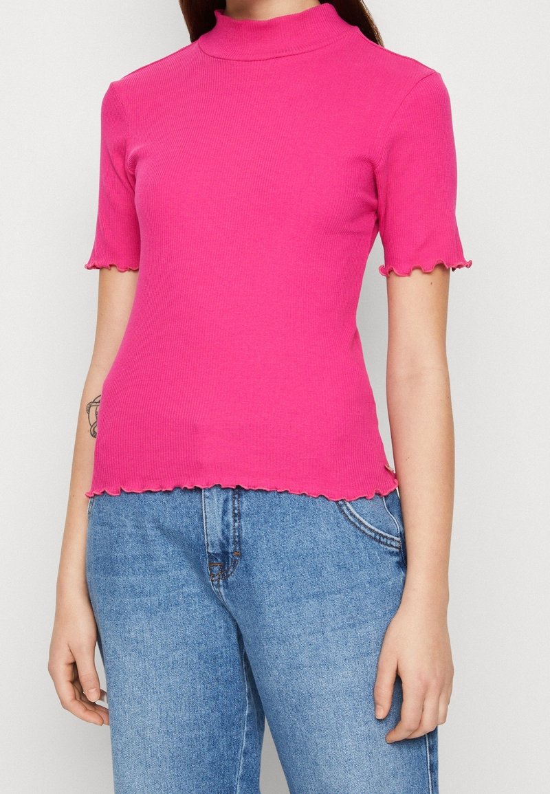 Haut rose ajusté à côtes avec un col montant et des manches courtes à bords festonnés, présentant un ourlet court et une texture douce. Porté avec des jeans bleus.