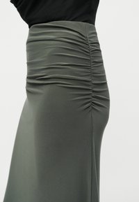 Φούστα χαμηλής μέσης σε ελιά πράσινο με λεπτομέρεια ραφής με ίσιωμα στη μία πλευρά, φορεμένη με μαύρο τοπ.