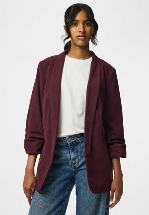 Jonge vrouw met donker haar, gekleed in een wit overhemd, een bordeauxblauwe blazer en een blauwe spijkerbroek, staand tegen een effen lichte achtergrond.