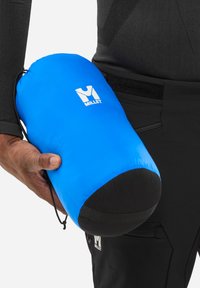 Personne tenant un sac de rangement Millet bleu avec une base noire, portant des vêtements de plein air noirs sur un fond blanc.