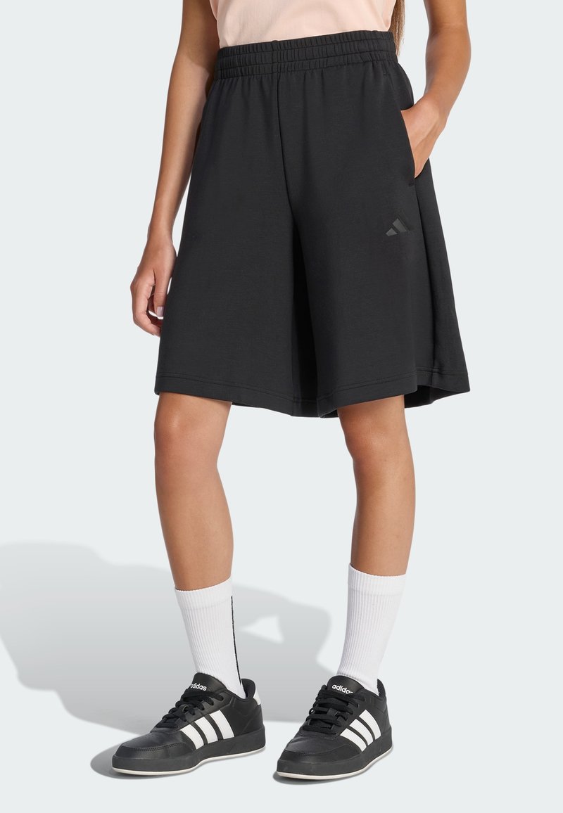 Pantaloncini sportivi neri con vita regolabile a coulisse, tasche laterali e un dettaglio del logo discreto; abbinati a scarpe da ginnastica nere e calzini bianchi.