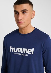 Mörkblå långärmad t-shirt i bomull med en framträdande vit "hummel"-logotyp och text, rund halslinning och slät textur.