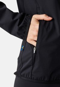 Zwarte softshell jas met een ritszak en een Zweedse vlagpatch. Soepele textuur en een afgestemde pasvorm rond de taille en mouwen.