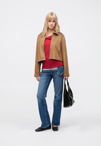 Brun ruskindsjakke i cropped model, rød top og blå jeans. Sort håndtaske og flade sko fuldender looket. Ensfarvet baggrund.