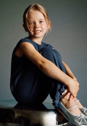 Barn iført en marineblå ribstrikket dragt med korte ærmer, siddende med krydsede ben på en metaloverflade. Fodtøj: sølvfarvede balletsko med sløjfer.