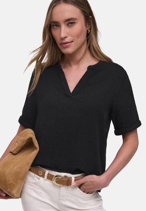 Femme portant un haut noir à manches courtes et col en V, un pantalon blanc avec une ceinture beige, tenant une pochette en daim beige sur un fond uni.