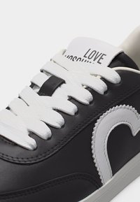 Μαύρο αθλητικό παπούτσι με λευκά κορδόνια, μια μεγάλη λευκή καρδιά στη πλευρά και μια λευκή ετικέτα στη γλώσσα με την επιγραφή "LOVE MOSCHINO".