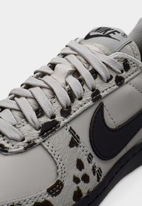 Svetlo sivo športno copat s poudarki iz usnja in vzorčastega konjskega dlaka. Ima črni logotip Nike, ravne vezalke in teksturirano površinsko podrobnost.