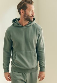 Zelena kapuljača veste od teksturiranog materijala, s prednjim džepom i rebrastim manžetama, u kombinaciji s odgovarajućim hlačama.