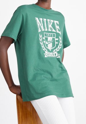 Osoba ubrana w zieloną koszulkę Nike z białym logo i napisem, siedząca na drewnianym krześle, w białych prążkowanych spodniach.