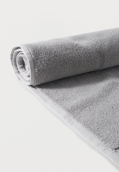 Πετσέτα από μικροΐνα σε ανοιχτό γκρι, μερικώς τυλιγμένη, παρουσιάζοντας μαλακή υφή με θηλιές και καθαρές άκρες πάνω σε λευκή επιφάνεια.