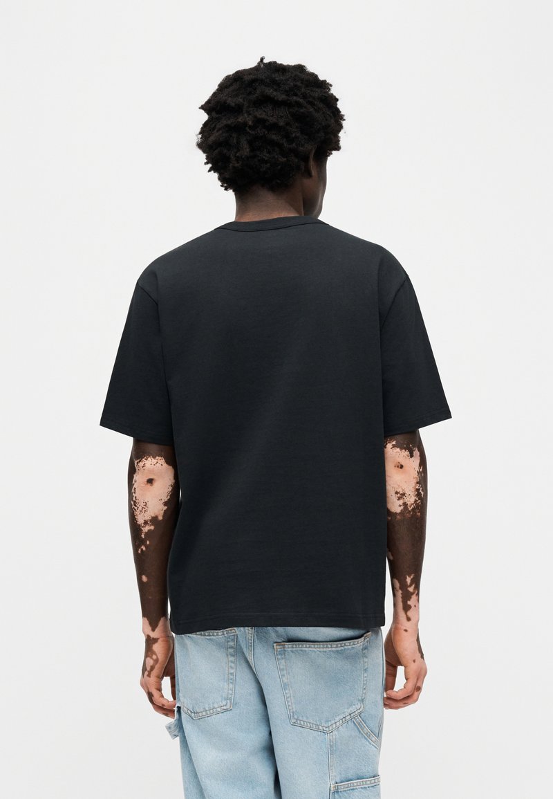 T-shirt nera in cotone con vestibilità morbida, collo rotondo e maniche corte. Indossata con pantaloni cargo in denim azzurro chiaro, che mostrano motivi unici sulle braccia.