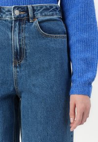 Jeans di denim azzurro chiaro con vita alta, tasca anteriore visibile e cuciture arancioni in contrasto. Un maglione blu testurizzato è parzialmente visibile.