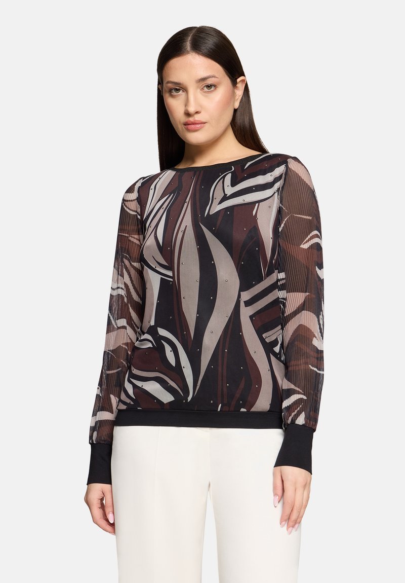 Femme portant un haut transparent à manches longues avec un motif abstrait noir, beige et marron et des poignets noirs, associé à un pantalon blanc.