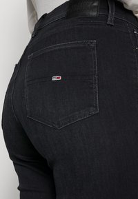 Pantalones vaqueros de mezclilla negra con un diseño ajustado, que cuentan con un bolsillo trasero adornado con un pequeño parche de logo en rojo, blanco y azul.