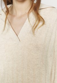 Beige stickad tröja med V-ringning, som har vertikal ribbning och en mjuk textur. Tyget ser lätt och något texturerat ut.