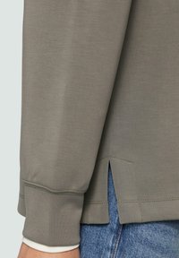 Maglia grigia chiaro a maniche lunghe con scollatura a girocollo, caratterizzata da una fessura laterale all'orlo e polsini a coste. Indossata sopra un paio di jeans blu.
