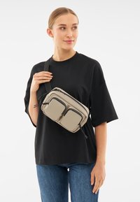 Beige axelremväska med två framfickor, justerbar rem, matt yta och svarta inslag. Bärs över en svart t-shirt och denimjeans.