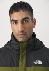 Giacca nera e verde con colletto alto, dotata di zip e chiusura a scatto. Il logo The North Face è ben visibile sul lato sinistro del petto.