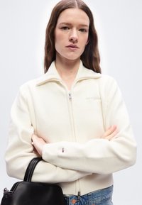 Νέα γυναίκα με μακριά καστανά μαλλιά, φορώντας ένα λευκό πουλόβερ με φερμουάρ και κρατώντας μια μαύρη δερμάτινη τσάντα, στέκεται μπροστά σε ένα απλό φόντο.