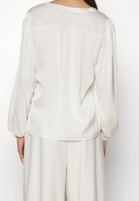 Weiße Bluse mit langen, puffärmeligen Ärmeln, glatter Textur und gerafftem Rückendetail. Leichter Stoff, leicht lockere Passform, runder Ausschnitt.