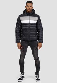 Chaqueta negra acolchada con rayas horizontales grises y blancas, con cremallera en el frente y capucha. Combinada con jeans negros rasgados y zapatillas negras.