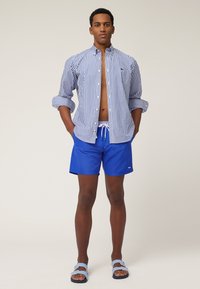 Gestreepte blauwe en witte blouse, blauwe zwemshorts en blauwe en witte gestreepte sandalen; heeft een relaxed fit en een zichtbaar merklogo.