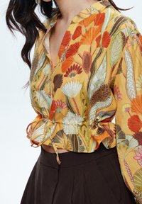 Blusa floreale in tessuto giallo con motivi multicolori di foglie e fiori. Maniche a sbuffo con dettaglio a coulisse in vita. Gonna marrone scuro.