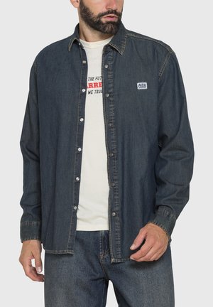 Uomo che indossa una camicia di denim scuro con bottoni sopra una t-shirt bianca con grafica e jeans blu, rivolto leggermente a sinistra con la mano vicino alla vita.